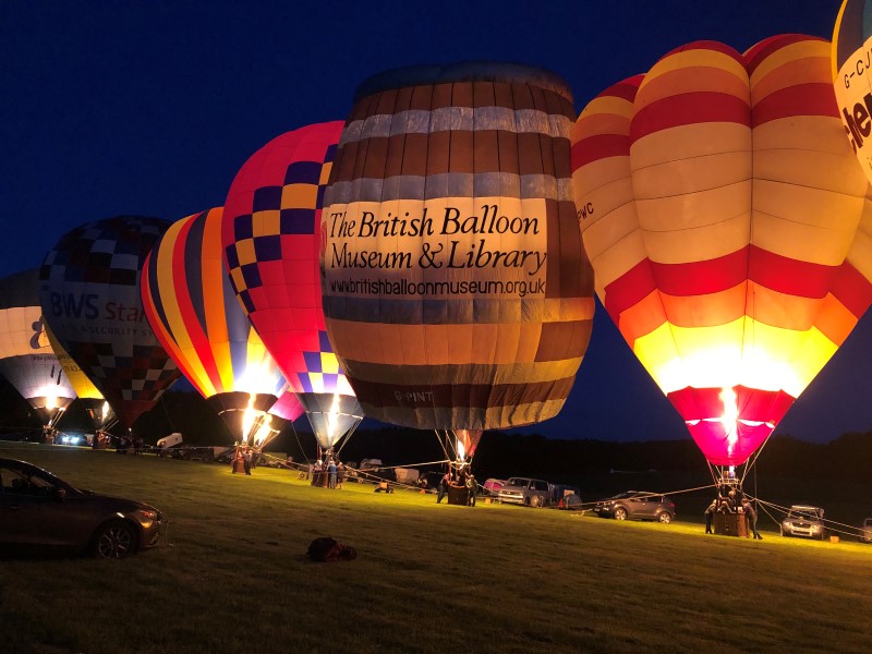 Balloon in a glow line up with other colourful balloons. The top half of the dark brown barrel with silver top and base, and four silver horizontal stripes representing the barrel rings, is lit up by the  burner flame, highlighting the BBM&L banner.